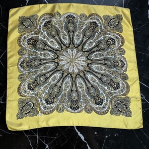 May include: A square scarf featuring a mustard yellow border and a central medallion design. The intricate pattern consists of black, white, and gold paisley motifs arranged in a radial, symmetrical floral-like formation across the fabric.