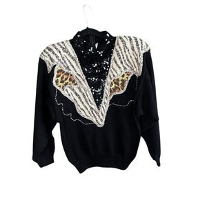 May include: Black sweater with a unique design. The collar and shoulder area feature a zebra print and leopard print pattern, with a sequined center. The sweater is hanging on a black hanger.