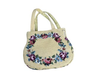 May include: A cream-colored handbag featuring a needlepoint floral wreath design. The bag has two top handles and a structured, rounded shape. The floral pattern consists of pink, purple, and blue flowers with green leaves.