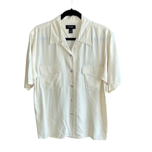 Puede incluir: Camisa crema de manga corta con dos bolsillos en el pecho. La camisa está colgada de una percha negra. La etiqueta dice "Silk House".