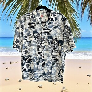 May include: A short-sleeved button-down shirt with a Hawaiian print. The shirt features a repeating pattern of Honolulu and Hawaii imagery in black and white. The shirt is hanging against a backdrop of a beach and palm trees.