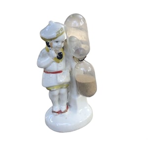 May include: A white porcelain figurine of a boy holding a small hourglass. The hourglass is made of glass and has sand inside. The figurine is standing on a white base.