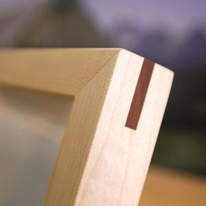 May include: A light-colored wooden picture frame corner featuring a miter joint reinforced with a contrasting dark brown rectangular wooden spline inlay. The frame has a smooth, matte finish and a clean, angled edge construction.