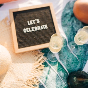 May include: A picnic blanket with a straw hat, two glasses of champagne, a bottle of champagne, and a letter board that says "Let's Celebrate".