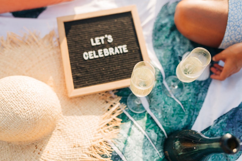 May include: A picnic blanket with a straw hat, two glasses of champagne, a bottle of champagne, and a letter board that says "Let's Celebrate".