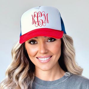 May include: A white and blue trucker hat with a red brim. The hat has a white front panel with red embroidered initials "KCM".