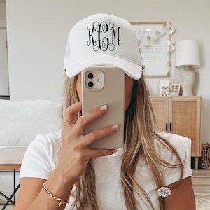 May include: White trucker hat with a black monogram. The hat has a curved brim and a mesh back. The monogram features the letters KCM in an elegant, cursive font. The hat is a stylish accessory.