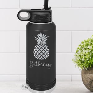 May include: A black stainless steel water bottle with a silver lid and a straw. The bottle has a pineapple design and the name "Bethany" engraved on it.