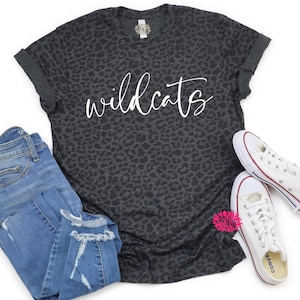 May include: A charcoal gray t-shirt with a leopard print pattern and the word "wildcats" in white script. The shirt is paired with blue jeans and white canvas sneakers with red accents. A pink flower adds a pop of color.