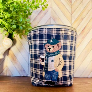 May include: A glass jar wrapped in a navy blue and white plaid fabric. The fabric features an embroidered bear wearing a green hat, scarf, and coat, holding a coffee cup. The bear is the focal point of the design.