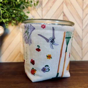 May include: A cylindrical fabric storage bin featuring a cream-colored background with a repeating pattern of gardening tools, including pruners, rakes, and various colorful flowers. A small white rectangular tag with black text is attached to the side.
