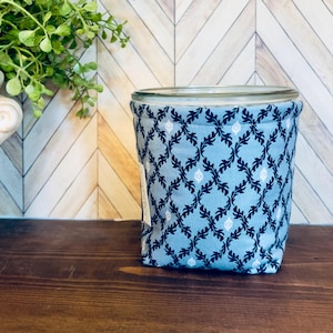 May include: A glass jar encased in a light blue fabric sleeve featuring a dark blue and white repeating damask-style pattern. The fabric is quilted and fits snugly around the cylindrical glass container, which has a clear rim at the top.