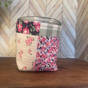 May include: A clear glass jar with a lid, wrapped in a patchwork fabric cozy. The fabric features floral patterns in pink, blue, and white, along with a gray and white gingham check. The jar sits on a dark wooden shelf.