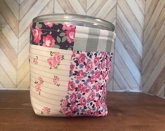 May include: A clear glass jar with a lid, wrapped in a patchwork fabric cozy. The fabric features floral patterns in pink, blue, and white, along with a gray and white gingham check. The jar sits on a dark wooden shelf.