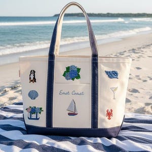 May include: A cream-colored canvas tote bag with navy blue trim and handles. The bag features embroidered designs including a dog, seashells, a sailboat, a martini glass, a lobster, and blue hydrangeas. The text East Coast is embroidered in blue.