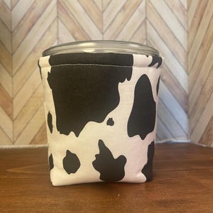 May include: A jar cozy with a black and white cow print design. The cozy is wrapped around a clear glass jar. The background is a wooden surface and a light brown chevron pattern.