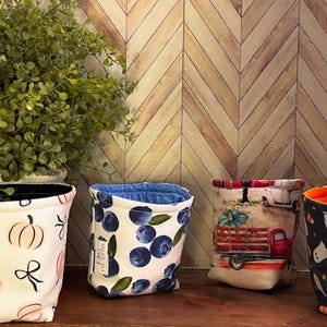 May include: Four fabric storage containers with different patterns. One has pumpkins and bows, another has blueberries, a third has a red truck, and the last has ghosts and candy corn. A plant in a ceramic pot is in the background.