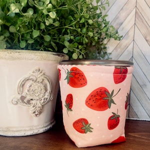 May include: A pink fabric drink sleeve featuring a pattern of red strawberries with green leaves and white polka dots. The sleeve is wrapped around a silver-rimmed container. A decorative white pot with a green plant is in the background.