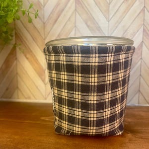 May include: A clear glass jar with a lid, wrapped in a black and cream plaid fabric. The jar sits on a wooden surface, with a light-colored, chevron-patterned background. The fabric wrap adds a decorative touch to the jar.