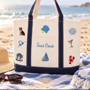 May include: A cream-colored canvas tote bag with navy blue trim and handles. The bag features embroidered icons including a dog, hydrangea, seashell, bikini, cocktail, lobster, sailboat, and a basket, with the text East Coast in blue script.