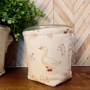 May include: A white fabric container with a duck illustration and floral accents. The container has a clear lid and is decorated with a repeating pattern of ducks and flowers in shades of beige and pink. The container is on a wooden surface.