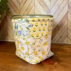 May include: A glass jar with a floral fabric cozy. The cozy features a patchwork design with yellow roses, blue flowers, and green leaves on a white background. The jar is sitting on a wooden surface.