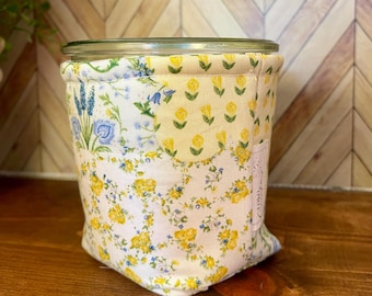 May include: A glass jar with a floral fabric cozy. The cozy features a patchwork design with yellow roses, blue flowers, and green leaves on a white background. The jar is sitting on a wooden surface.