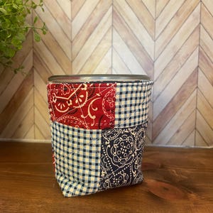 May include: A patchwork drink cozy featuring red paisley, navy paisley, and blue and white gingham fabrics. The cozy is designed to fit around a glass or can, providing insulation and a decorative touch. The design is quilted and stitched.