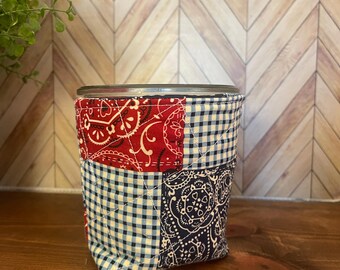 May include: A patchwork drink cozy featuring red paisley, navy paisley, and blue and white gingham fabrics. The cozy is designed to fit around a glass or can, providing insulation and a decorative touch. The design is quilted and stitched.