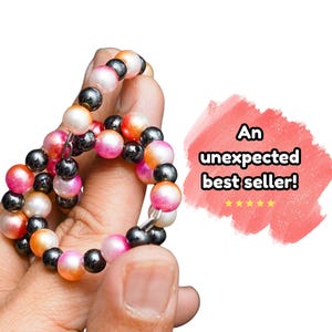 May include: A beaded bracelet featuring alternating spherical beads in metallic black and a gradient of pink, orange, and white. A coral-colored graphic element to the right displays the text "An unexpected best seller!" with five yellow stars.