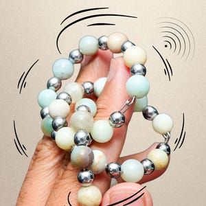 May include: A beaded bracelet from the Orbi Loops Luxe Series featuring alternating round light blue, cream, and tan stone beads interspersed with smaller, reflective silver-toned metallic spheres, arranged in a continuous circular loop pattern.