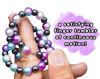 May include: A circular beaded loop featuring metallic dark gray, pearlescent white, vibrant purple, and bright blue beads strung on a flexible wire. The text "a satisfying finger tumbler of continuous motion!" is printed on a purple background.