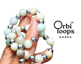 May include: A beaded bracelet featuring alternating round light green, beige, and tan stone beads interspersed with small, reflective silver-toned metal spheres. The text Orbi Loops with five stars is printed in black to the right of the jewelry.