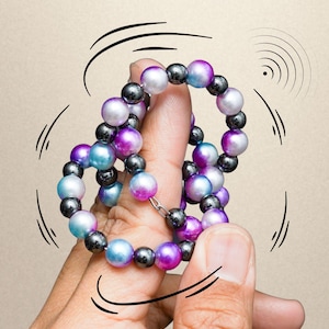 Peut inclure: Un bracelet de perles composé de sphères métalliques sombres alternant avec des perles lisses multicolores dans des tons de violet, bleu et blanc. Le texte Orbi Loops Essential Series est imprimé en haut, avec des lignes de mouvement graphiques.