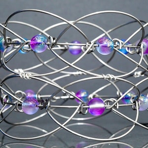 May include: A silver wire bracelet with iridescent purple and blue beads. The bracelet is made of multiple strands of wire that are woven together to create a complex design.