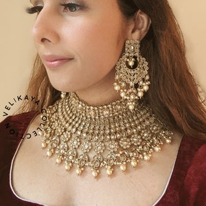 May include: A woman wearing a maroon velvet blouse with a gold and pearl necklace and matching earrings. The necklace is a multi-layered design with a large central pendant. The earrings are long and dangle with a similar design to the necklace. The text "VELIKAYA COLLECTION" is visible at the top of the image.