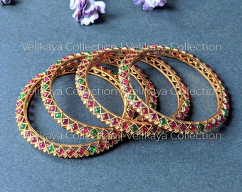 May include: Four gold bangles with red and green gemstones in a geometric design. The bangles are on a dark grey surface, with purple flowers in the background. The text "Velikaya Collection" is visible.