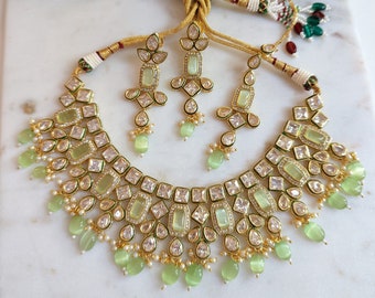 Op de afbeelding: Een gouden ketting en oorbellen set met groene en witte stenen. De ketting heeft een vierkant ontwerp met een rij groene stenen en een rij witte stenen. De oorbellen zijn hangende oorbellen met een groene steen en een witte steen.