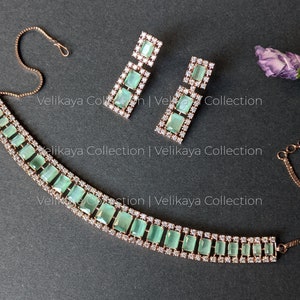 May include: A rose gold necklace and earrings set with a row of light green square-shaped stones surrounded by clear rhinestones. The necklace has a delicate chain.