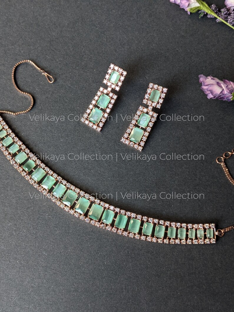 May include: A rose gold necklace and earrings set with a row of light green square-shaped stones surrounded by clear rhinestones. The necklace has a delicate chain.