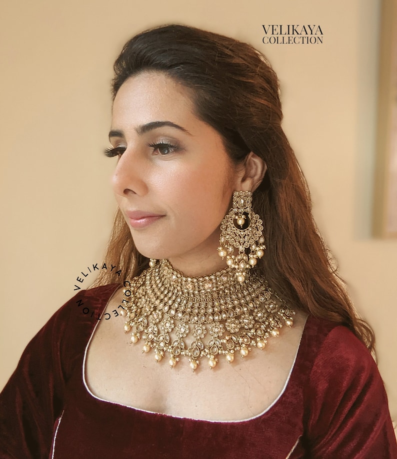 May include: A woman wearing a maroon velvet blouse with a gold and pearl necklace and matching earrings. The necklace is a multi-layered design with a large central pendant. The earrings are long and dangle with a similar design to the necklace. The text "VELIKAYA COLLECTION" is visible at the top of the image.