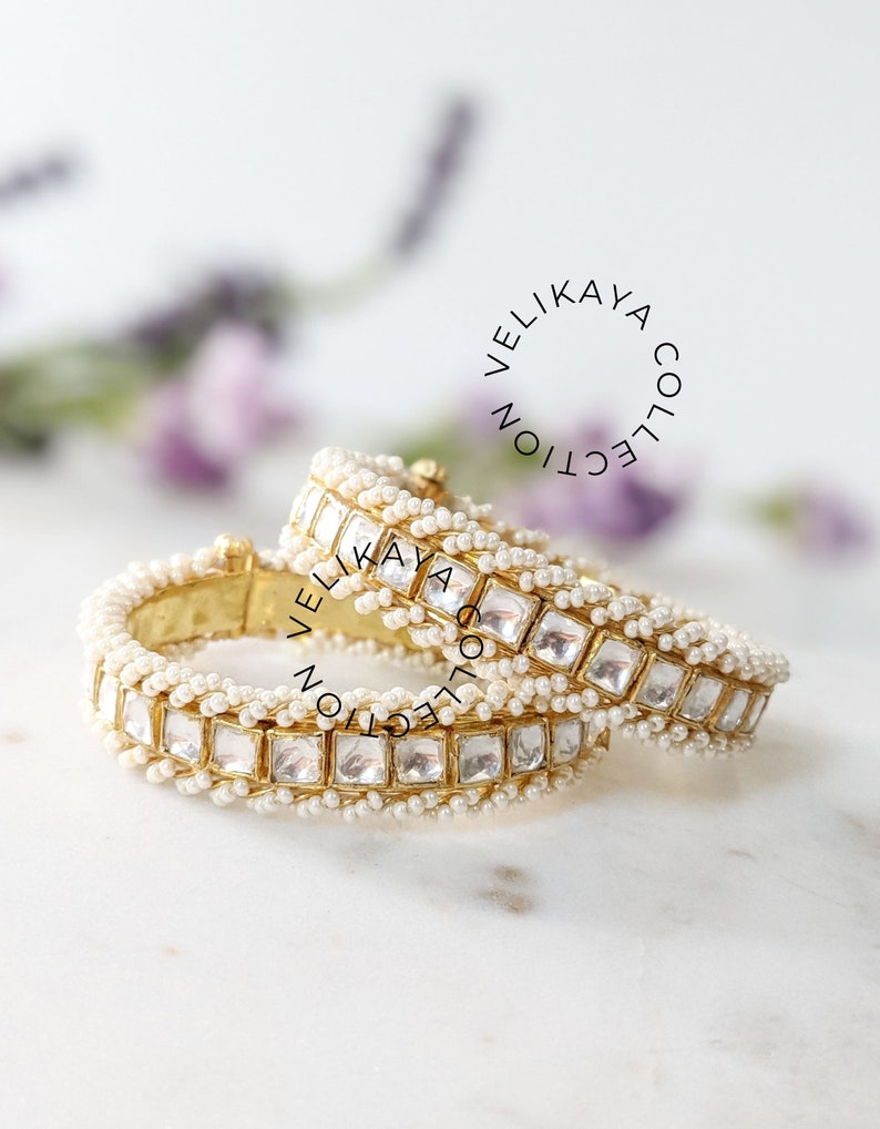 May include: Two gold bangles with white pearl and clear square stone embellishments. The bangles are stacked on top of each other. The text "Velikaya Collection" is visible in the background.