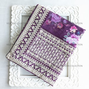 Op de afbeelding: Een paarse en witte geborduurde stof met een bloemenprint. De stof is gevouwen en wordt getoond in een wit houten frame.