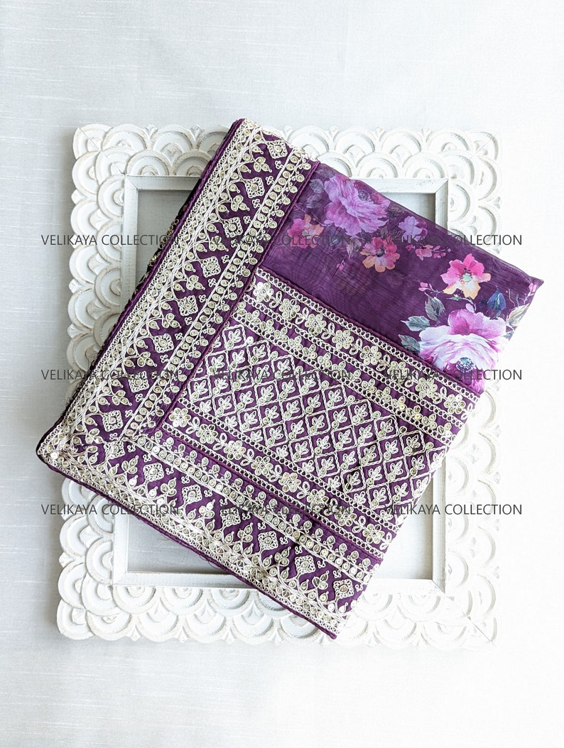 Op de afbeelding: Een paarse en witte geborduurde stof met een bloemenprint. De stof is gevouwen en wordt getoond in een wit houten frame.
