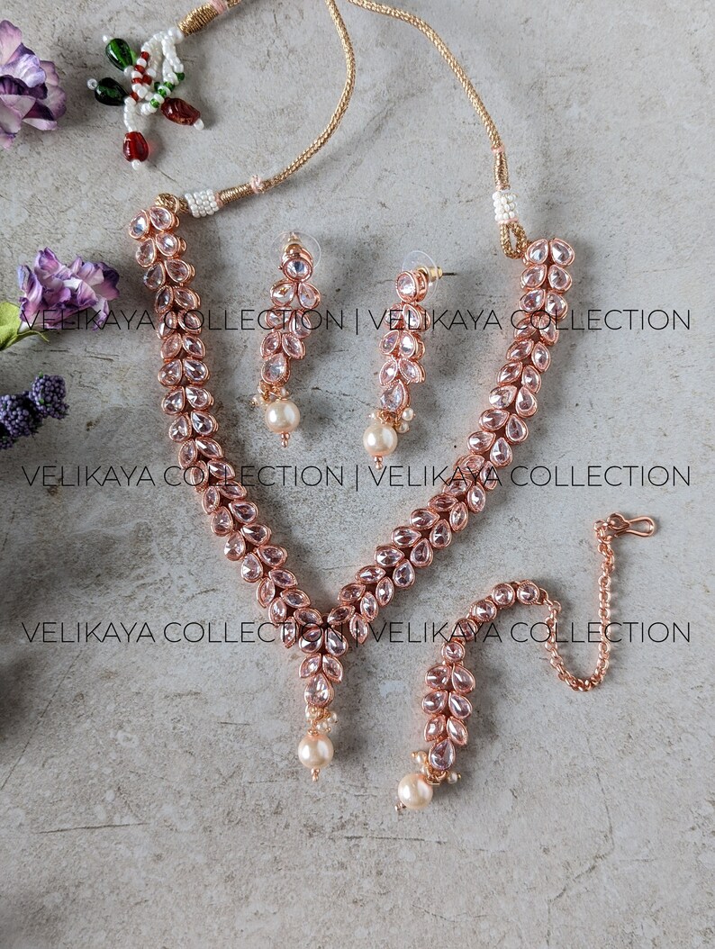 May include: A rose gold necklace set with a delicate chain, pink crystal teardrop embellishments, and pearl accents. The set includes a necklace, earrings, and a bracelet.