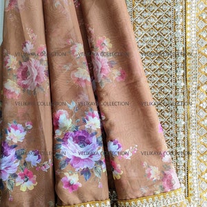 Op de afbeelding: Een bruine, doorschijnende stof met een bloemenprint en een gouden rand met witte pailletten. De stof heeft de tekst "VELIKAYA COLLECTION" erop gedrukt.
