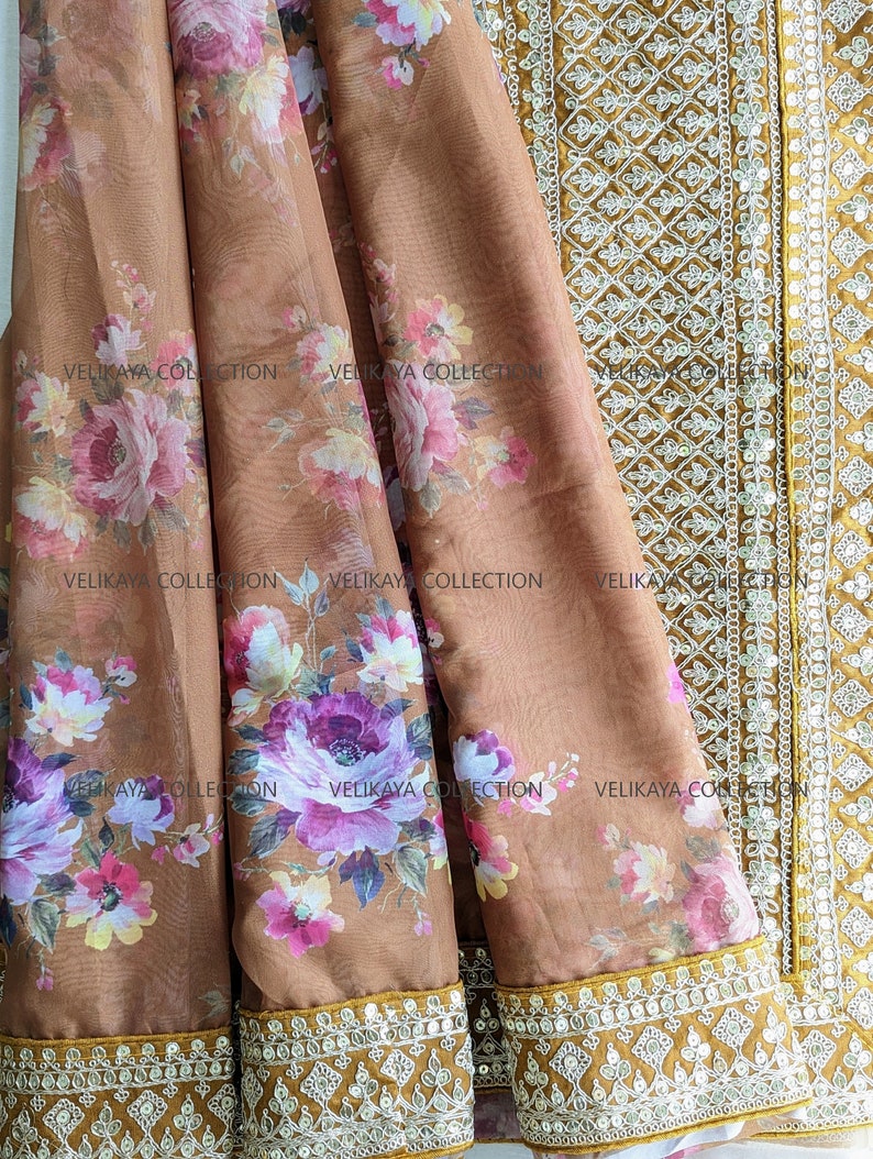 Op de afbeelding: Een bruine, doorschijnende stof met een bloemenprint en een gouden rand met witte pailletten. De stof heeft de tekst "VELIKAYA COLLECTION" erop gedrukt.