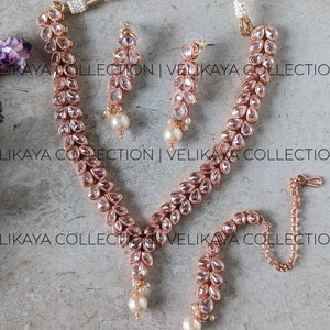 May include: A rose gold necklace and earrings set with a matching maang tikka. The jewelry is adorned with clear, faceted stones and pearl accents.