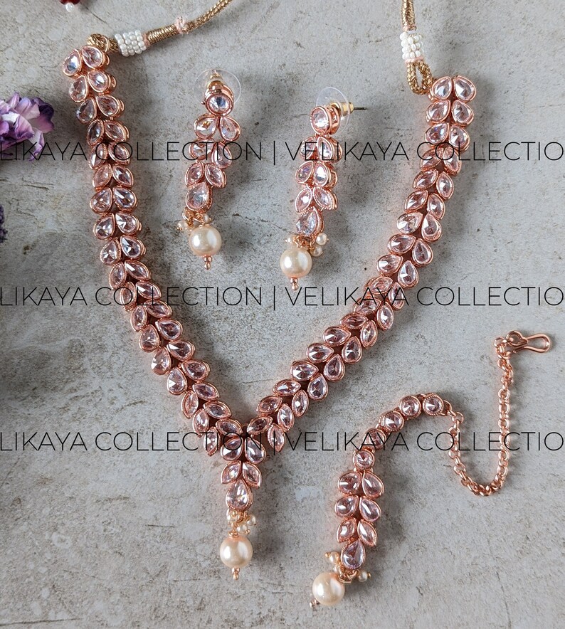 May include: A rose gold necklace and earrings set with a matching maang tikka. The jewelry is adorned with clear, faceted stones and pearl accents.