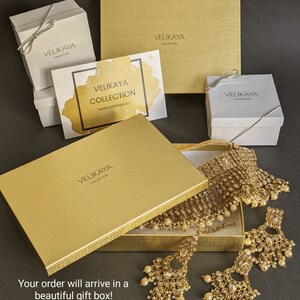 May include: A gold jewelry box with a gold necklace, earrings, and a matching headpiece. The box is open and the jewelry is displayed inside. The box has the text "Velikaya Collection" printed on it. The text "Your order will arrive in a beautiful gift box!" is printed on the bottom of the image.